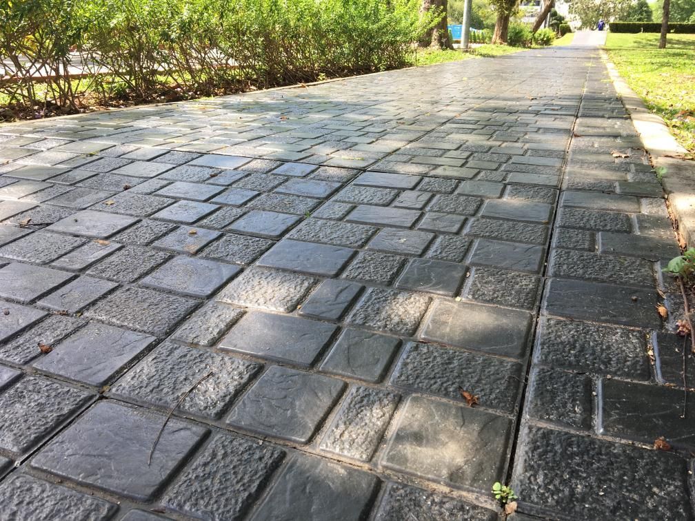 Dark gray brick-patterned walkway in a park, with green grass and bushes nearby.