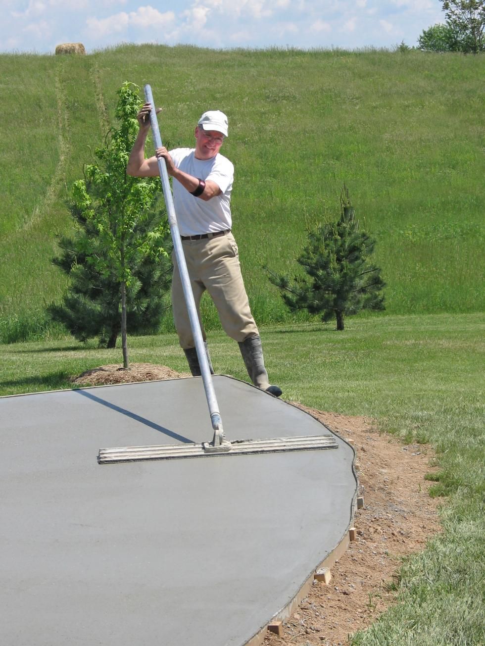 Man using a concrete leveler to smooth a freshly poured concrete patio in a grassy yard.