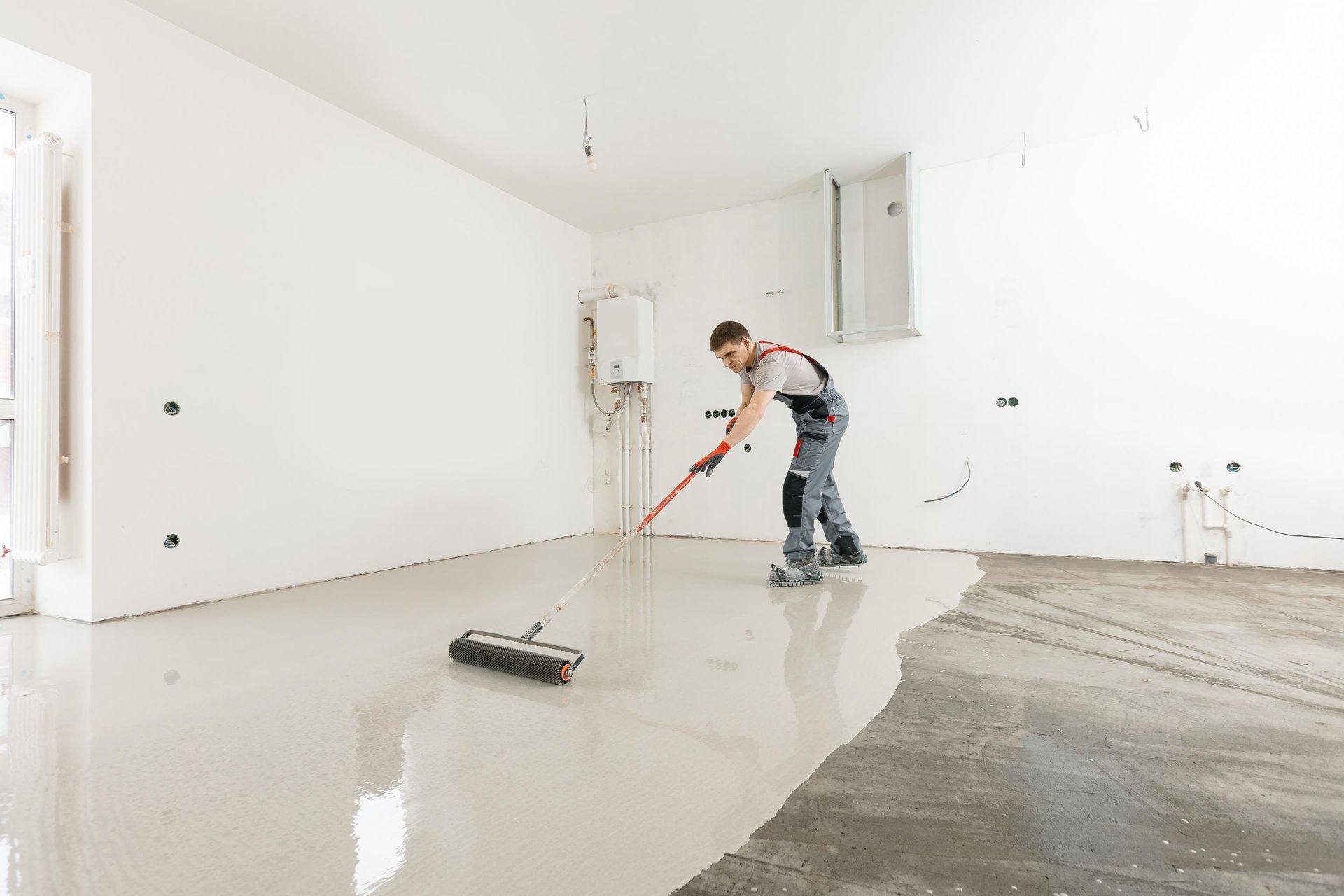 Man applying self-leveling concrete to a floor in a white-walled room.