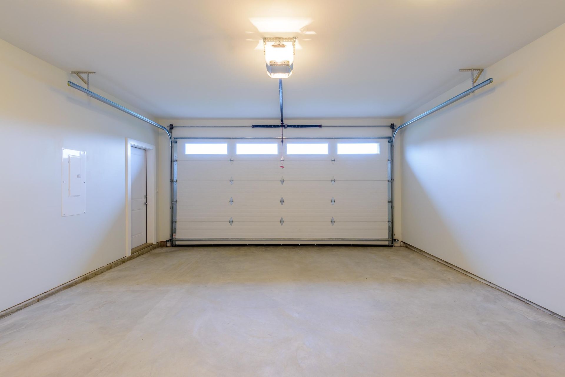 Empty, light-filled garage with a closed white door, concrete floor, and an overhead light fixture.