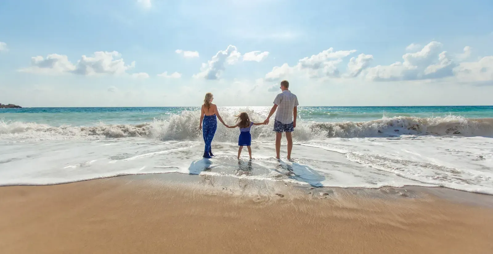 Famiglia di tre persone che si tengono per mano, in piedi su una spiaggia sabbiosa, guardando il mare.