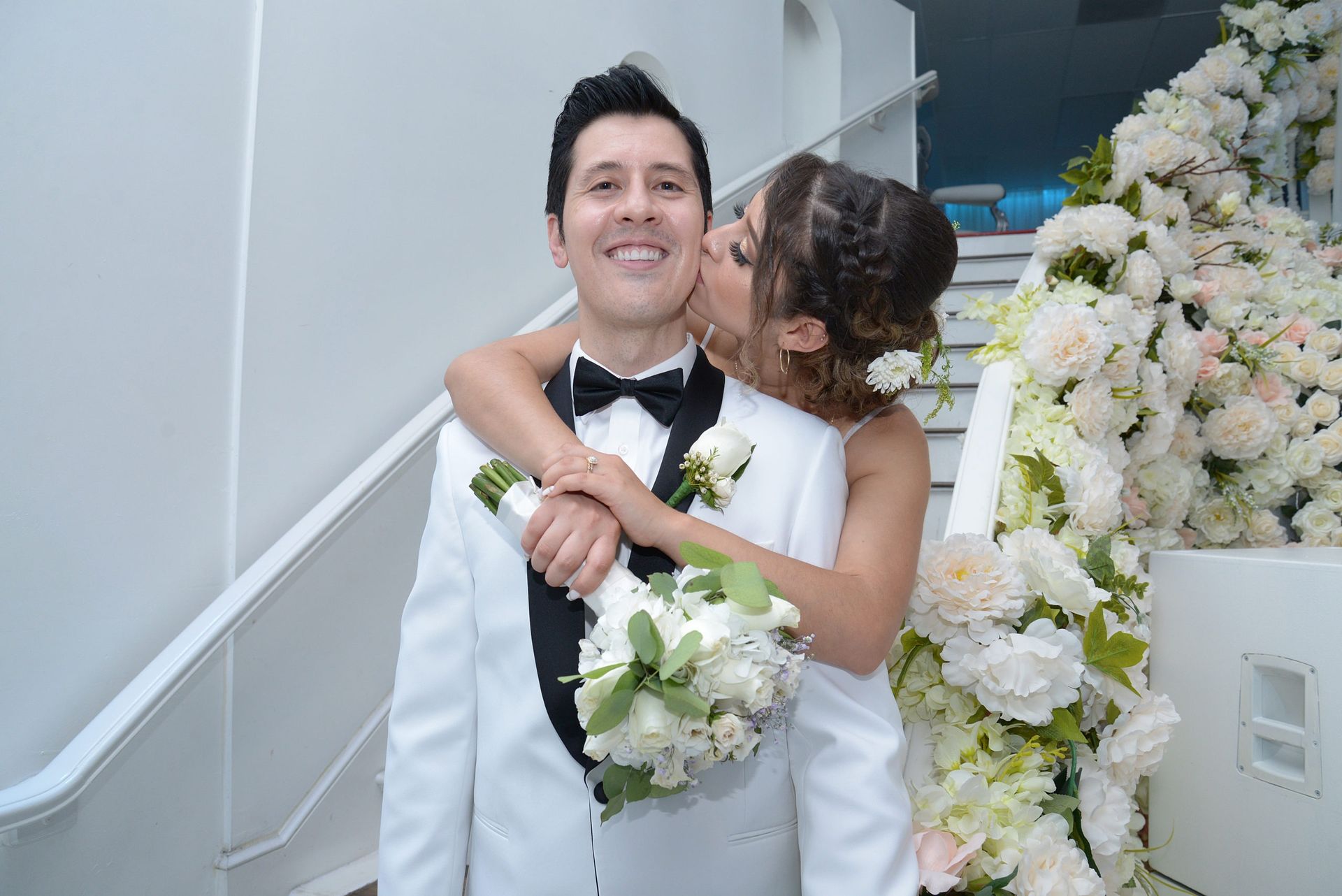 A bride is kissing a groom on the cheek while holding a bouquet of flowers.