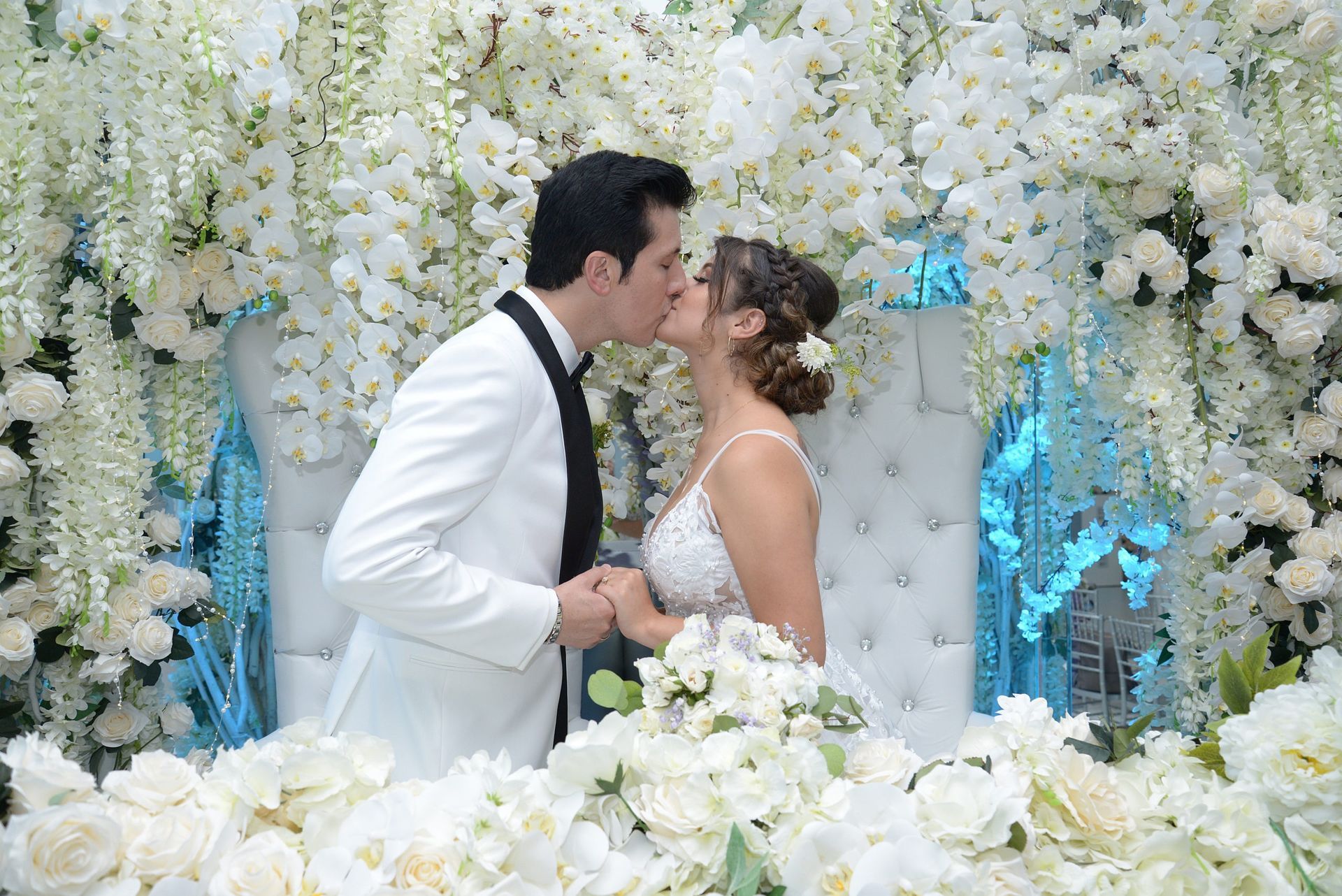 A bride and groom are kissing in front of a wall of white flowers.