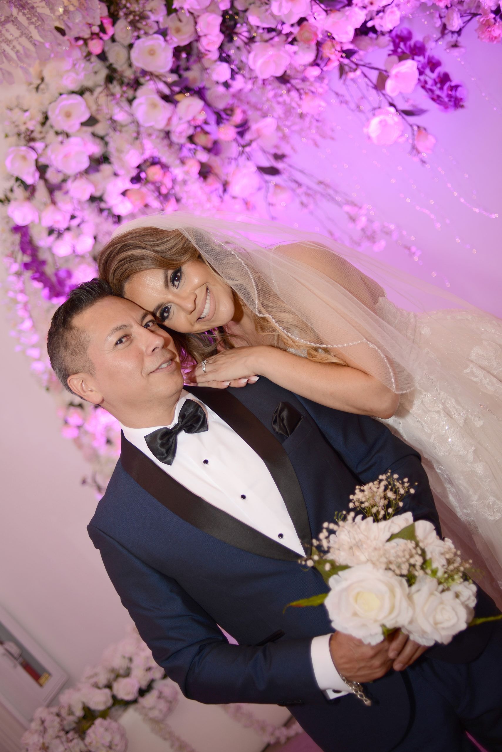 A bride and groom are posing for a picture in front of a wall of flowers.