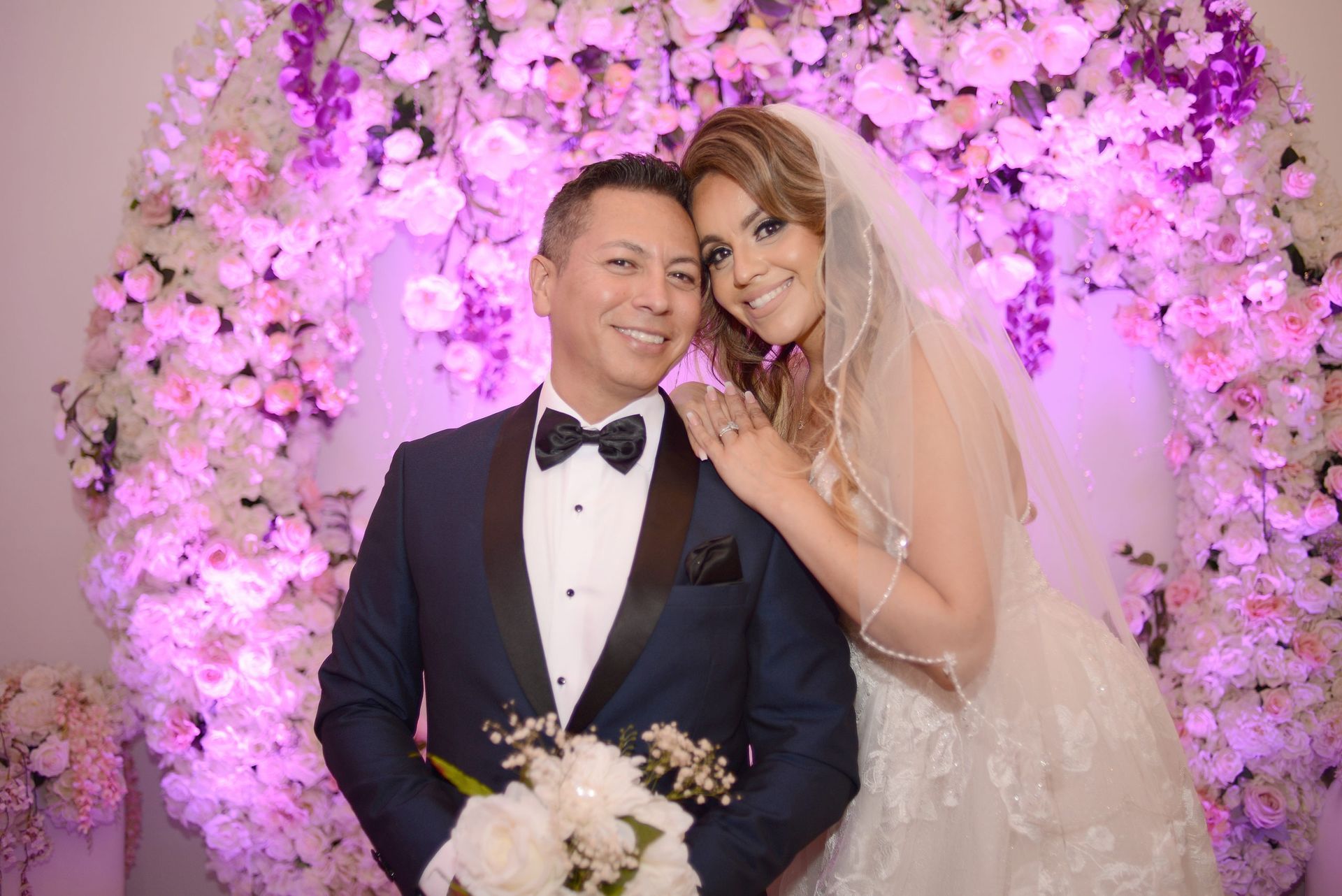 A bride and groom are posing for a picture in front of a wall of flowers.