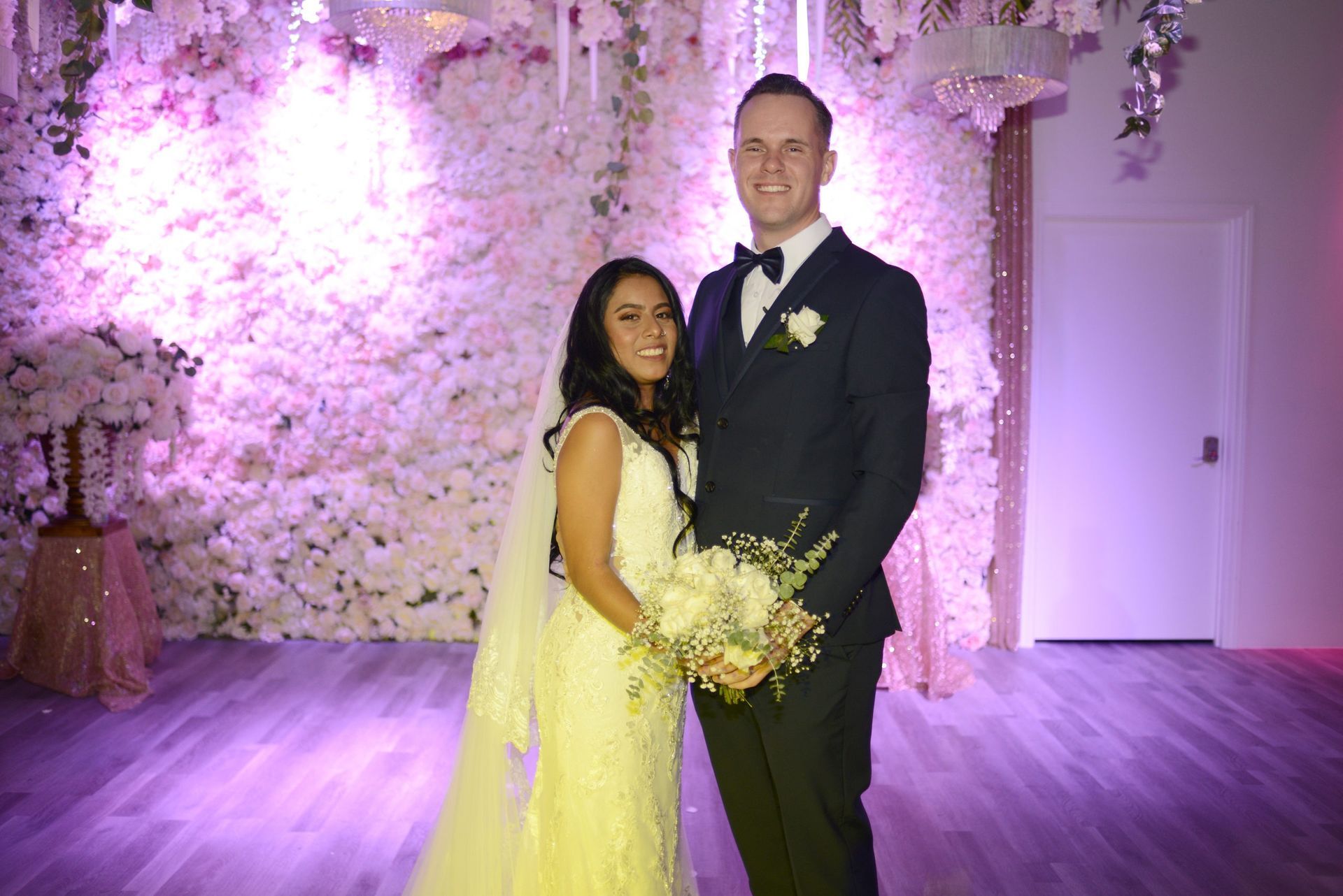 A bride and groom are posing for a picture in front of a wall of flowers.