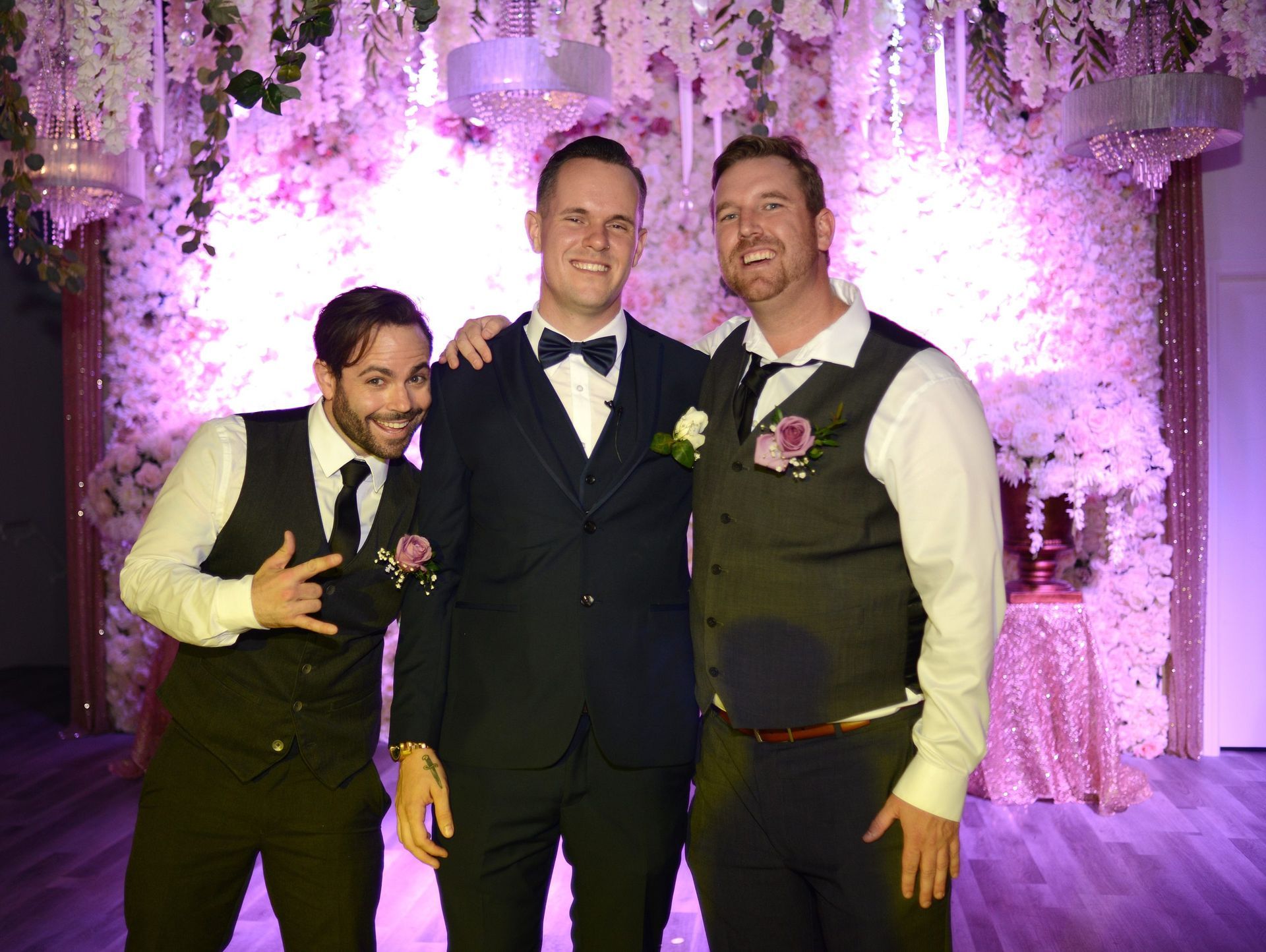 Three men are posing for a picture in front of a wall of flowers.