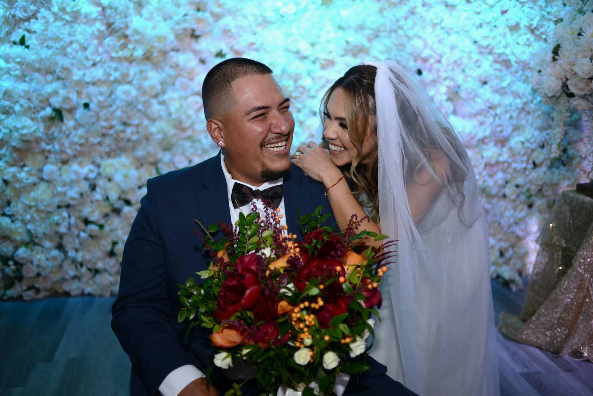 A bride and groom are posing for a picture in front of a wall of flowers.