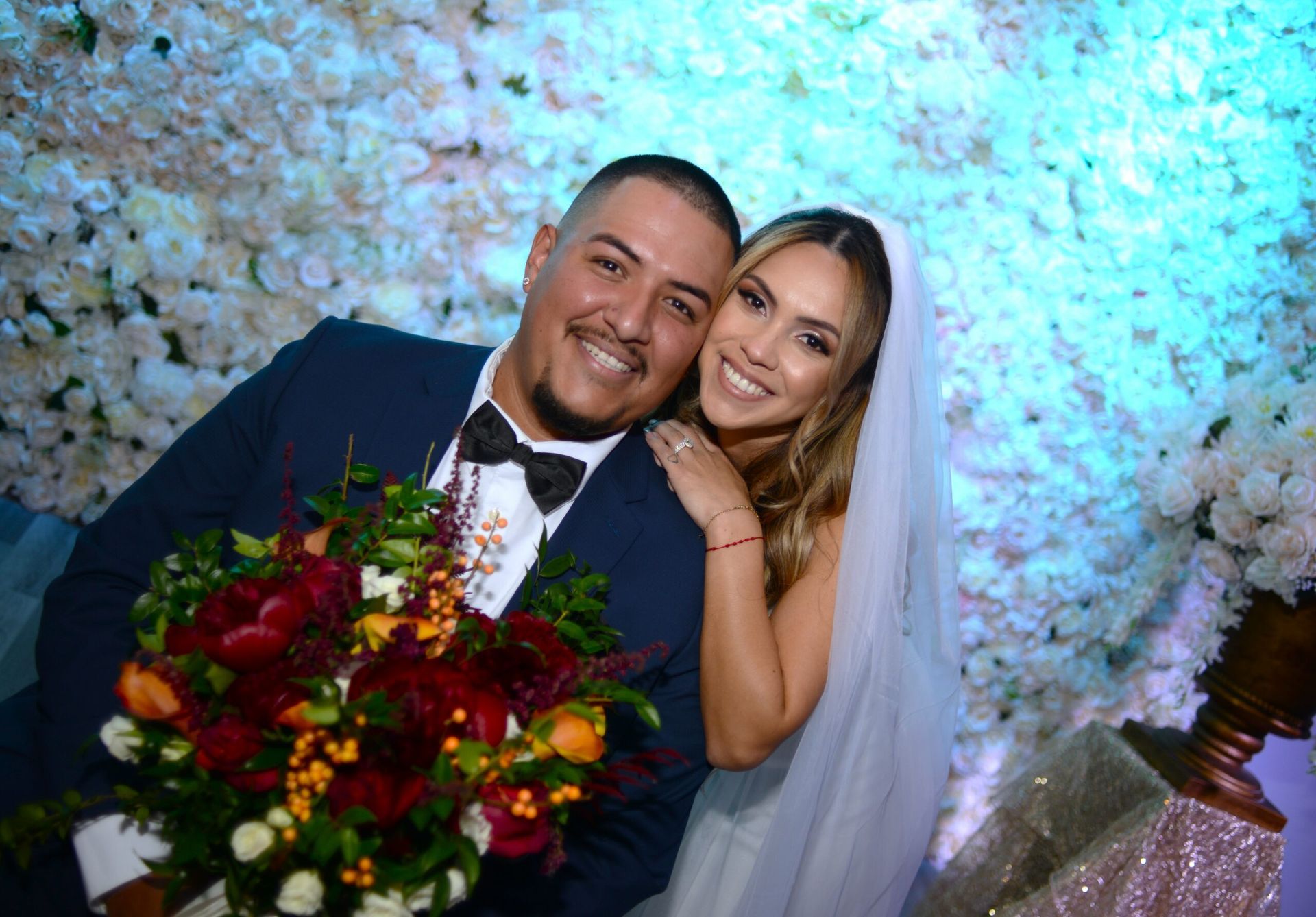 A bride and groom are posing for a picture in front of a wall of flowers.