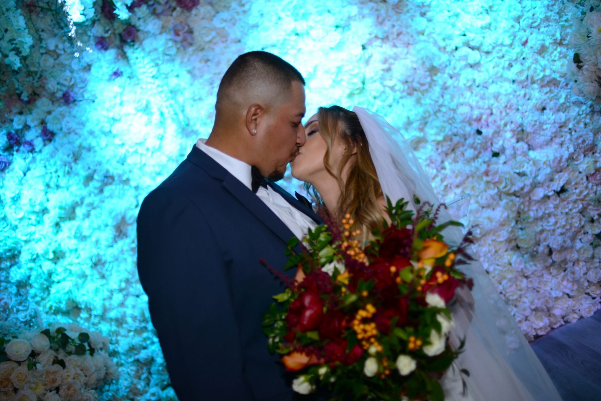 A bride and groom kissing in front of a wall of flowers.