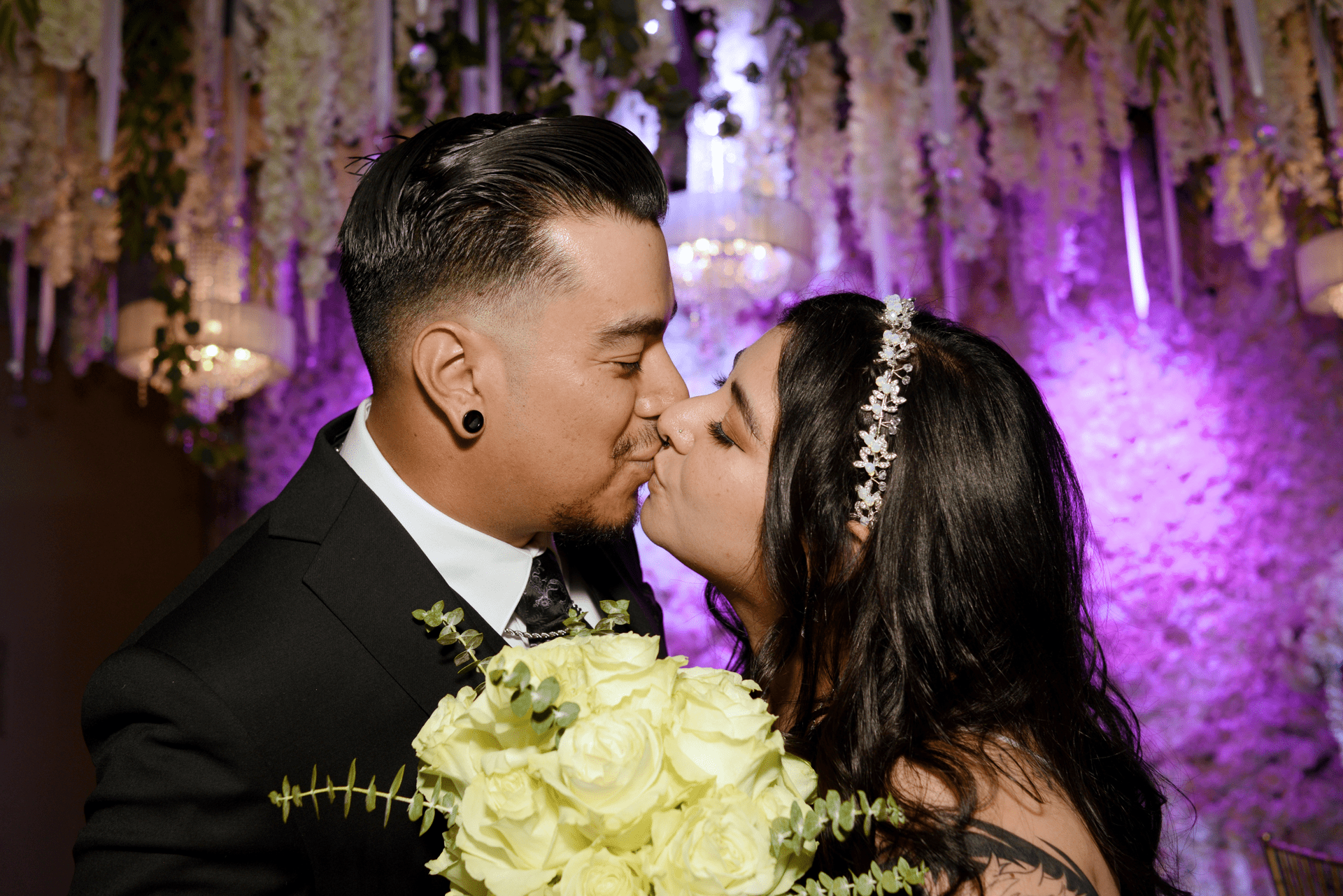 A bride and groom are kissing in front of a chandelier at their wedding reception.