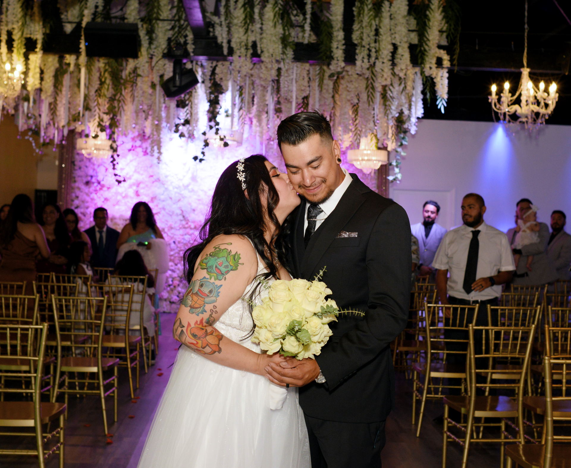 A bride and groom are kissing at their wedding ceremony in front of a crowd of people.