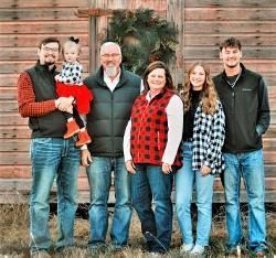 Family of six poses in front of a wooden structure; some wear plaid, smiling.