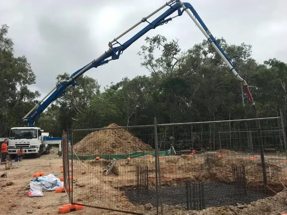 Concrete being poured at a construction site by a blue boom truck; pile of dirt and rebar visible.