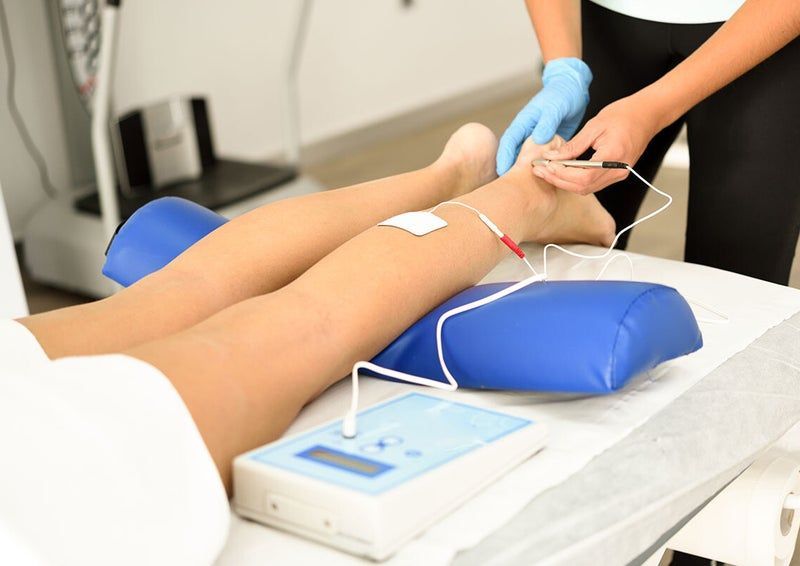 Persona sottoposta a elettroterapia sulle gambe, con elettrodi e un apparecchio in un ambiente medico.