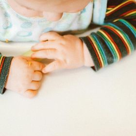 Le mani del bambino poggiano su una superficie bianca, indossa una maglietta a righe colorate su uno sfondo scuro.