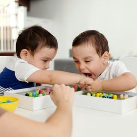 Due bambini piccoli seduti a un tavolo, uno cerca di afferrare i giocattoli di legno dall'altro, che sta piangendo.