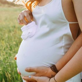 Persona incinta in abito bianco che tiene in mano le scarpette del bambino, con le mani del partner sulla pancia