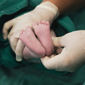 Mani nei guanti che tengono i piedi di un neonato, appoggiate su un telo chirurgico verde.