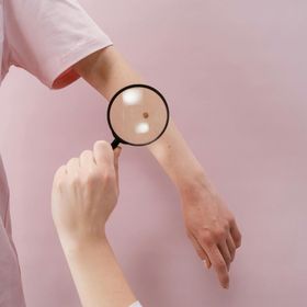 Persona che usa una lente d'ingrandimento per esaminare un neo sul braccio. Sfondo rosa.