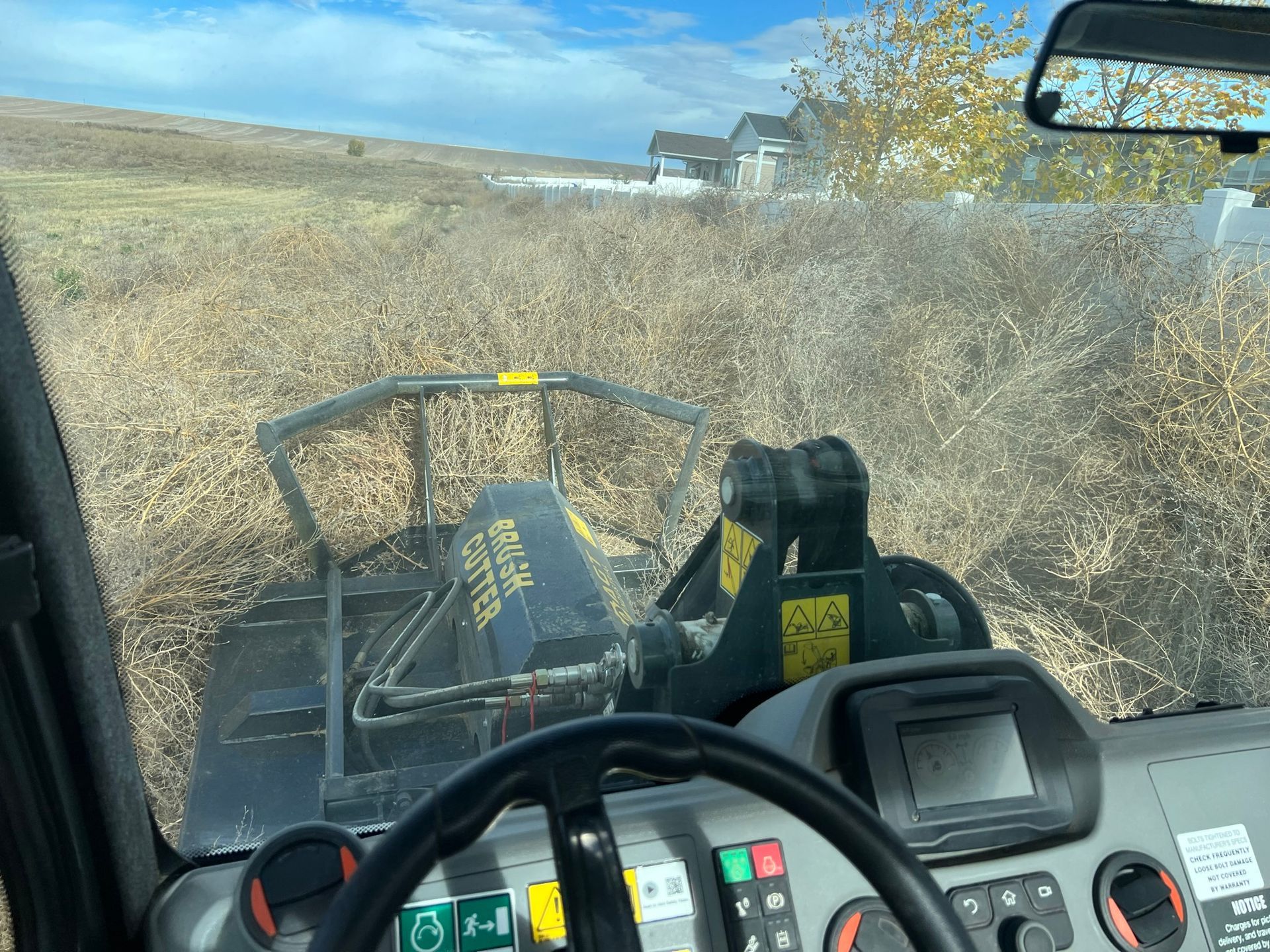 View from inside a machine clearing tall, dry weeds near a residential area.