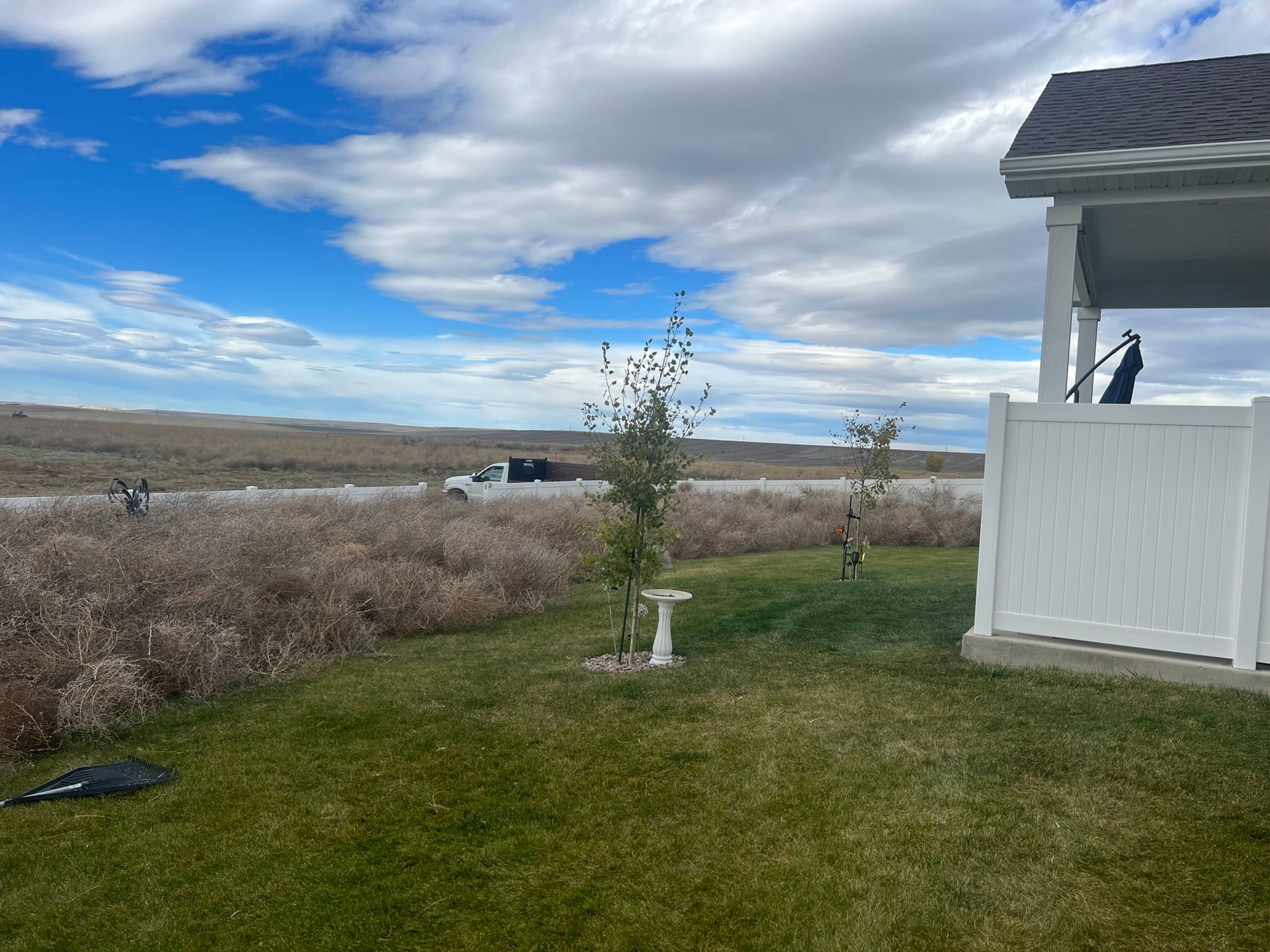 A backyard lawn with a white fence and an open field in the distance under a cloudy sky.