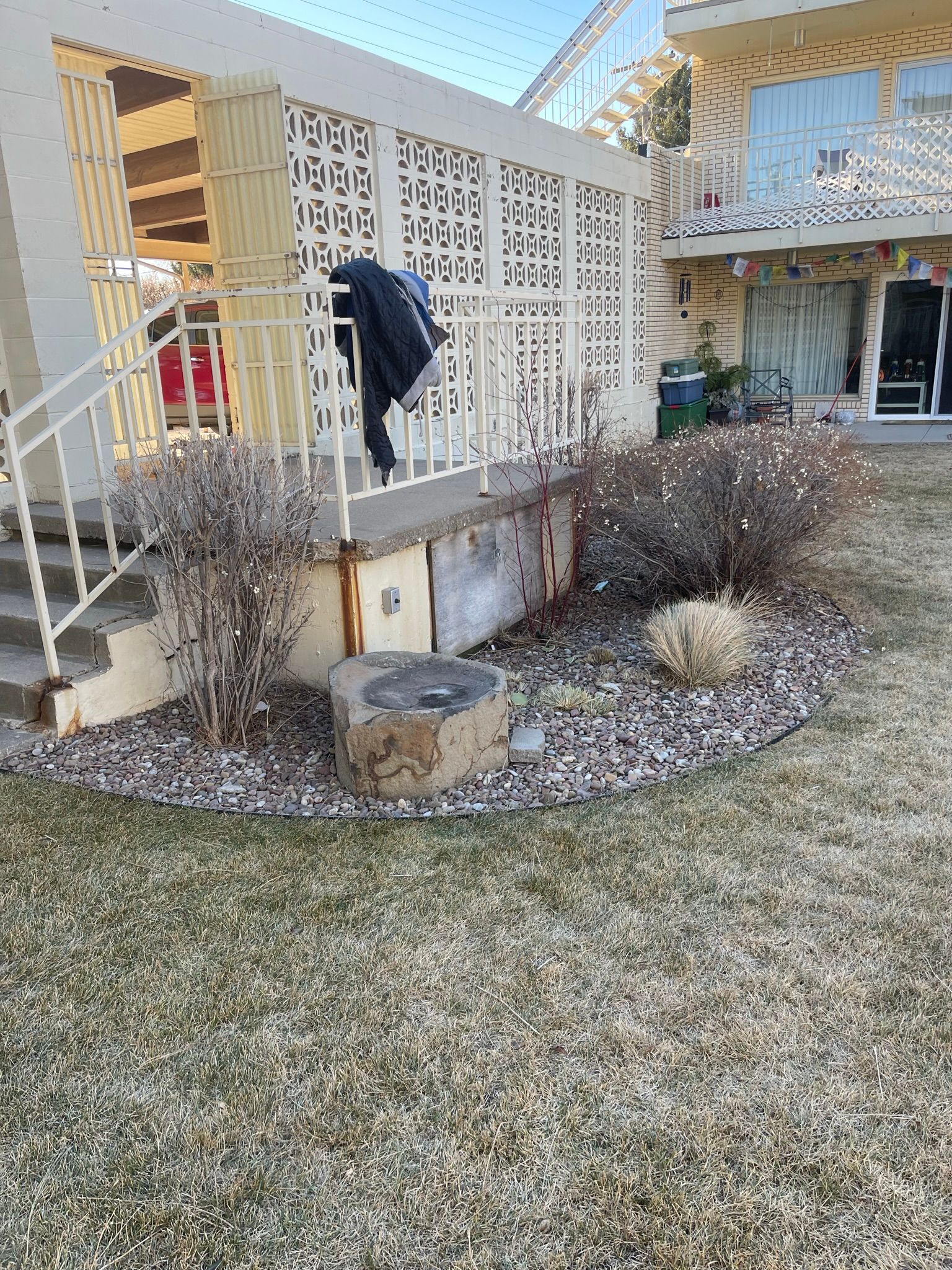A person wearing dark clothes tending to a front yard garden bed with dry brush and a concrete front porch.