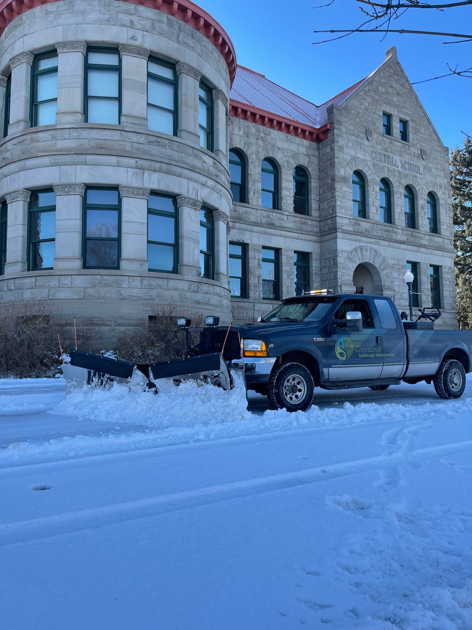 Snowplow truck clearing snow in front of a stone building with a curved turret on a winter day.
