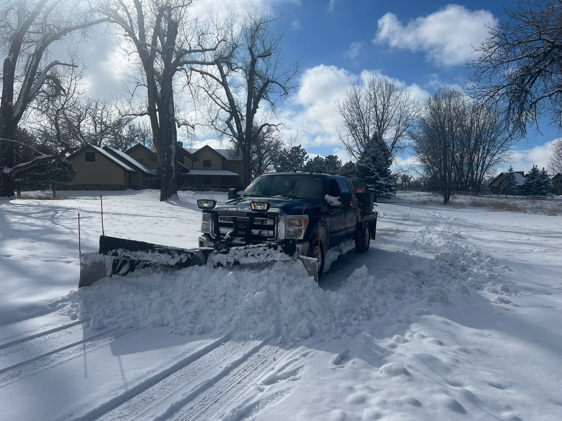 Blue pickup truck with snowplow clearing snow on a snowy day; houses and trees in the background.