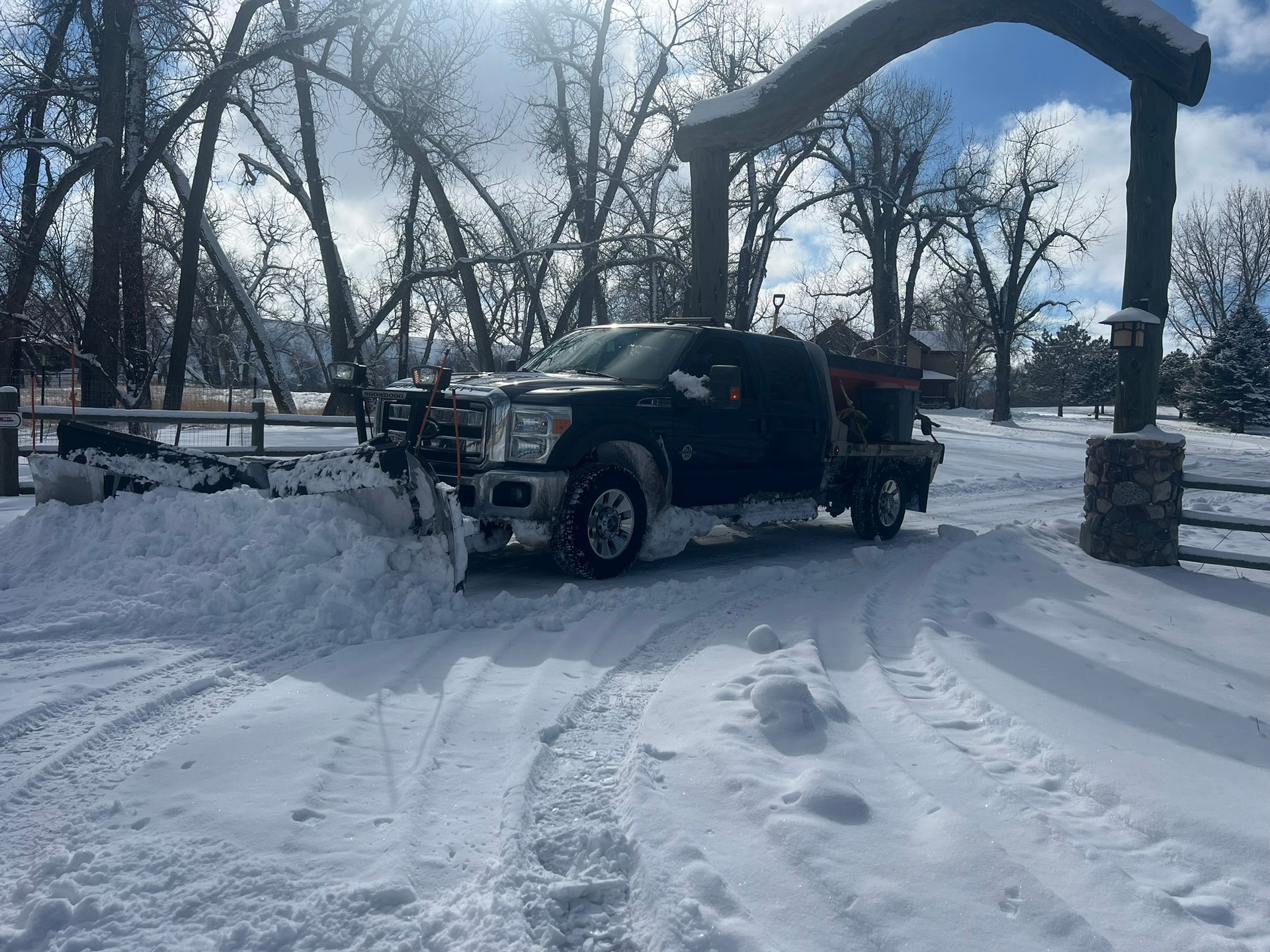 A black pickup truck with a snowplow clearing snow from a driveway on a sunny winter day.