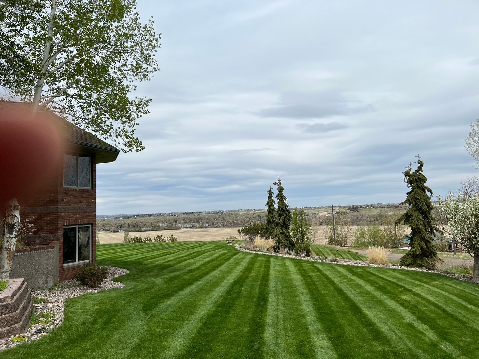 Lush green lawn with stripes, brick house, trees, and a cloudy sky. Landscape view.