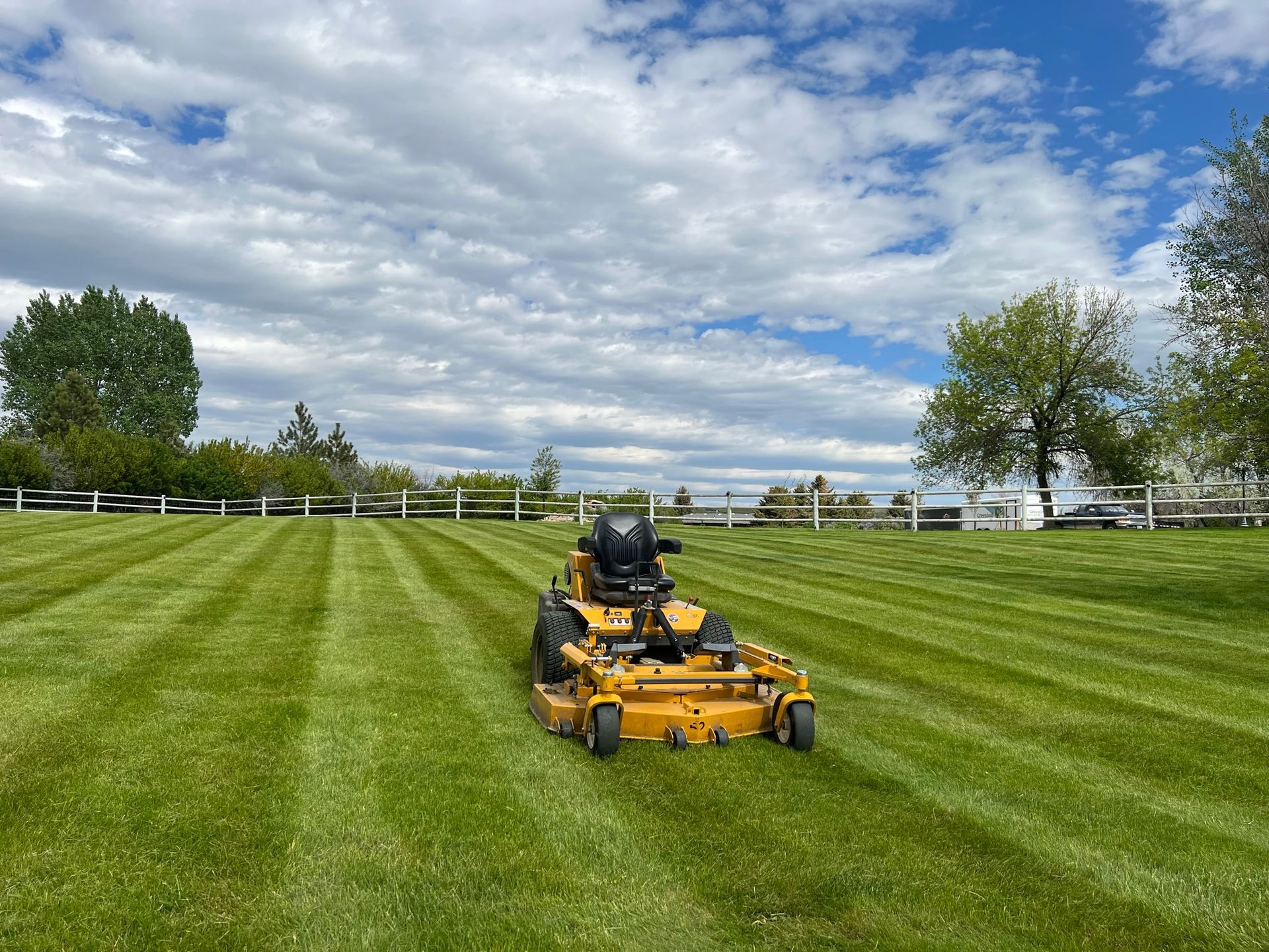 Yellow riding lawnmower cutting stripes in a green lawn on a sunny day, white fence in the background.
