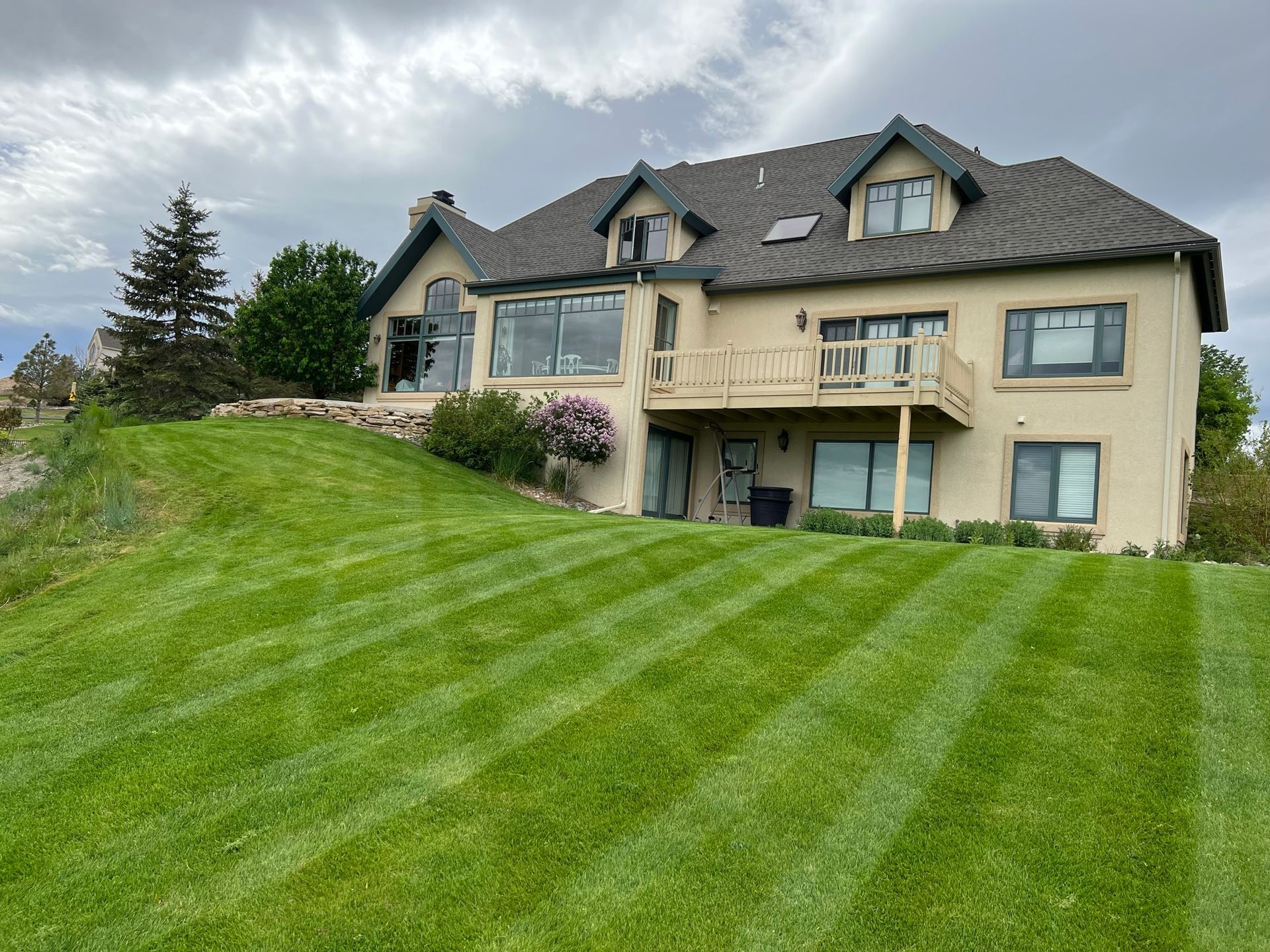 Two-story tan house on a hill with a well-manicured green lawn. Overcast sky.