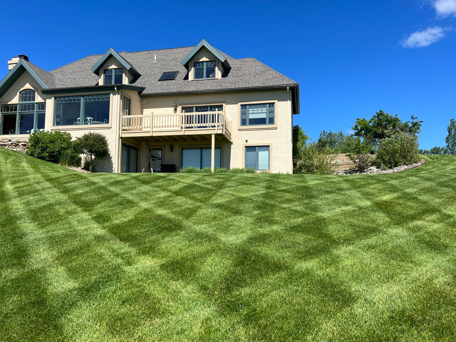 Lush green lawn mowed in a checkered pattern, in front of a beige house with blue sky backdrop.