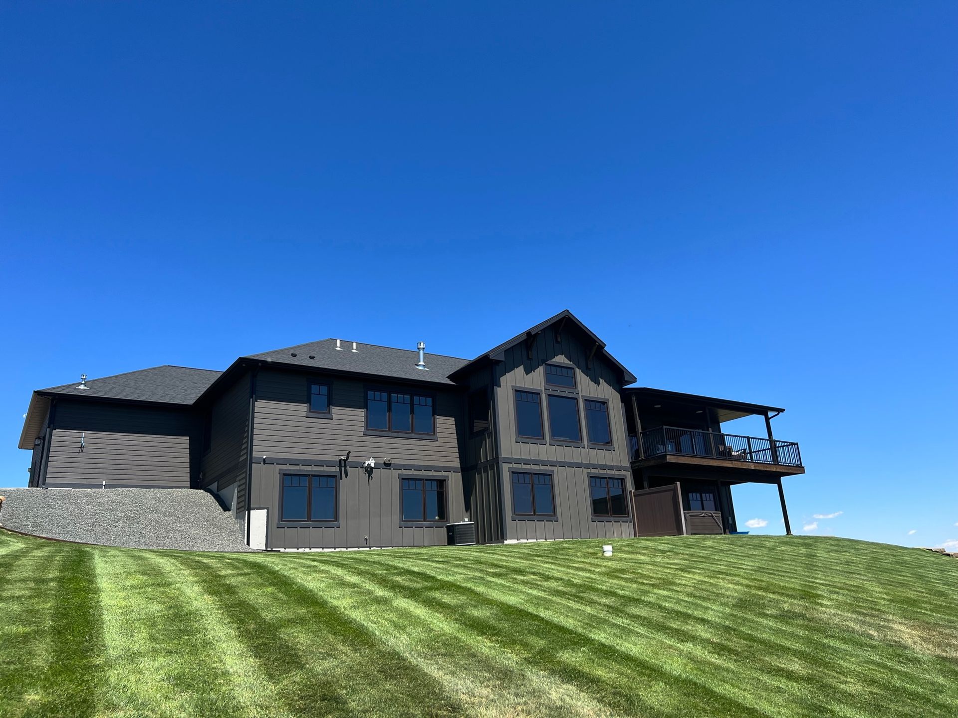 Modern gray house on a grassy hill under a clear blue sky, with a deck and dark roof.