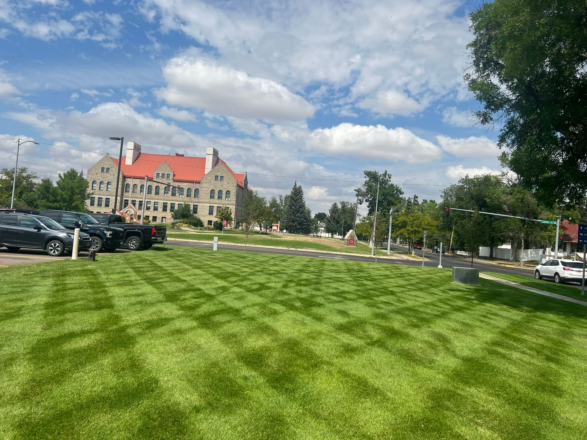 Lush green lawn mowed in a checkerboard pattern, with a large stone building in the background under a blue sky.