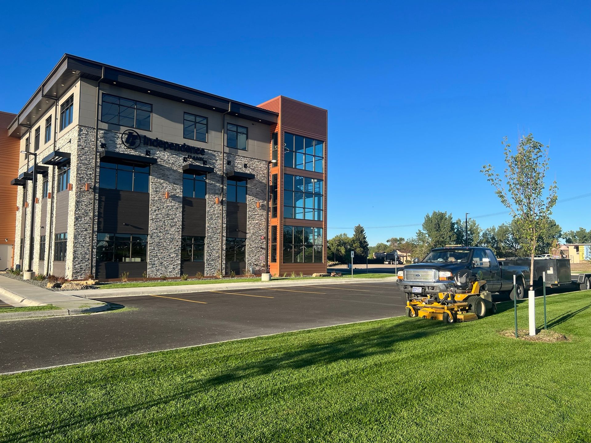 Modern building with stone and glass facade. Lawn mower on grass next to a trailer. Blue sky.