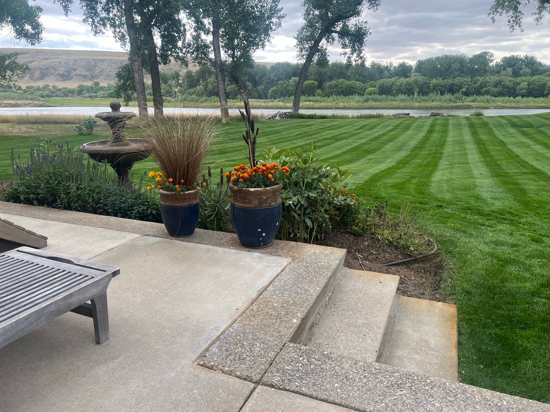 Patio overlooking a river with a fountain, potted plants, and a striped green lawn.