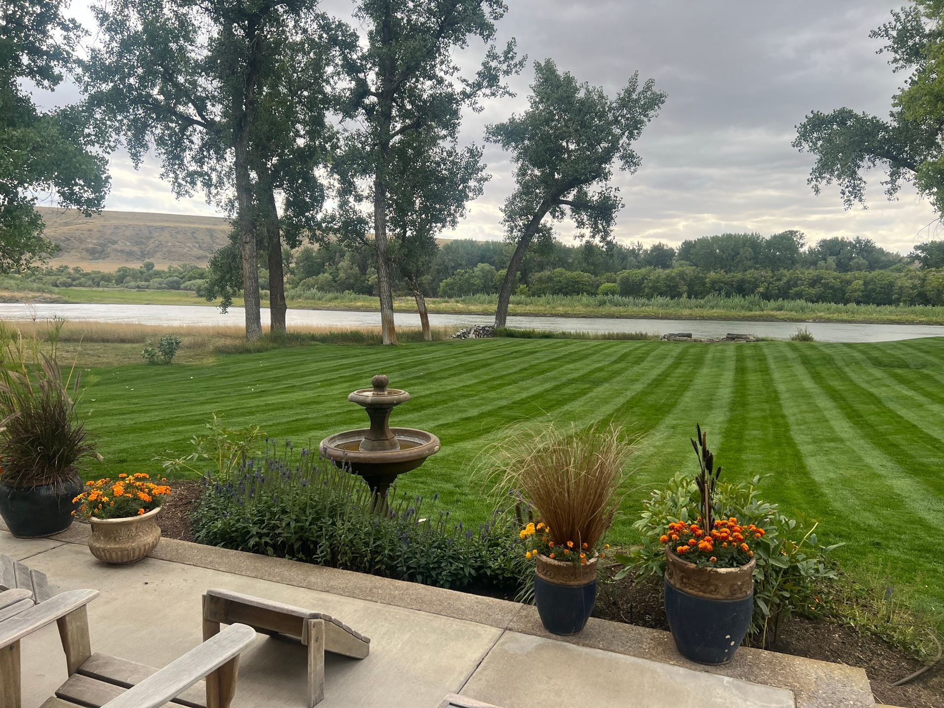 Lawn with a striped pattern, a fountain, and potted flowers overlooks a river under a cloudy sky.