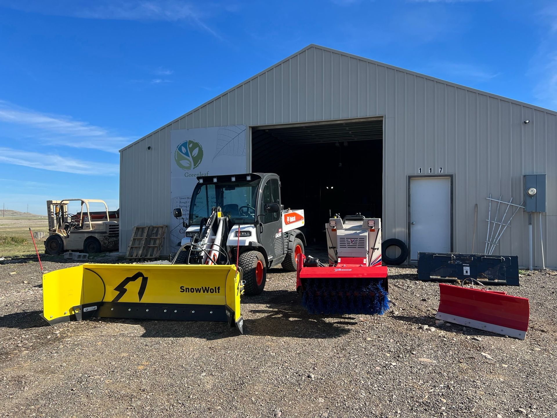 A Bobcat skid-steer loader with snow removal attachments outside a metal building on a sunny day.