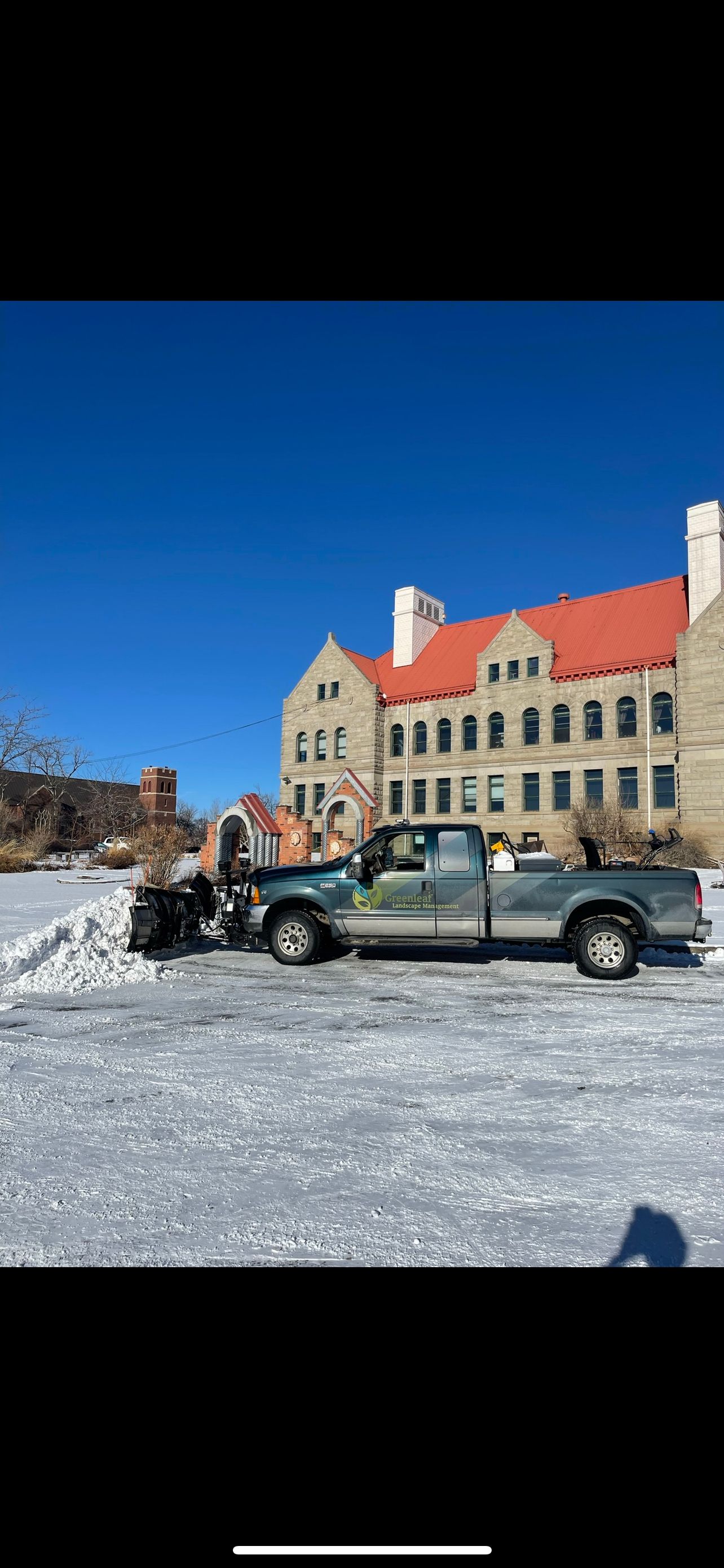 A snowplow truck clearing snow in front of a large brick building with a red roof on a sunny day.