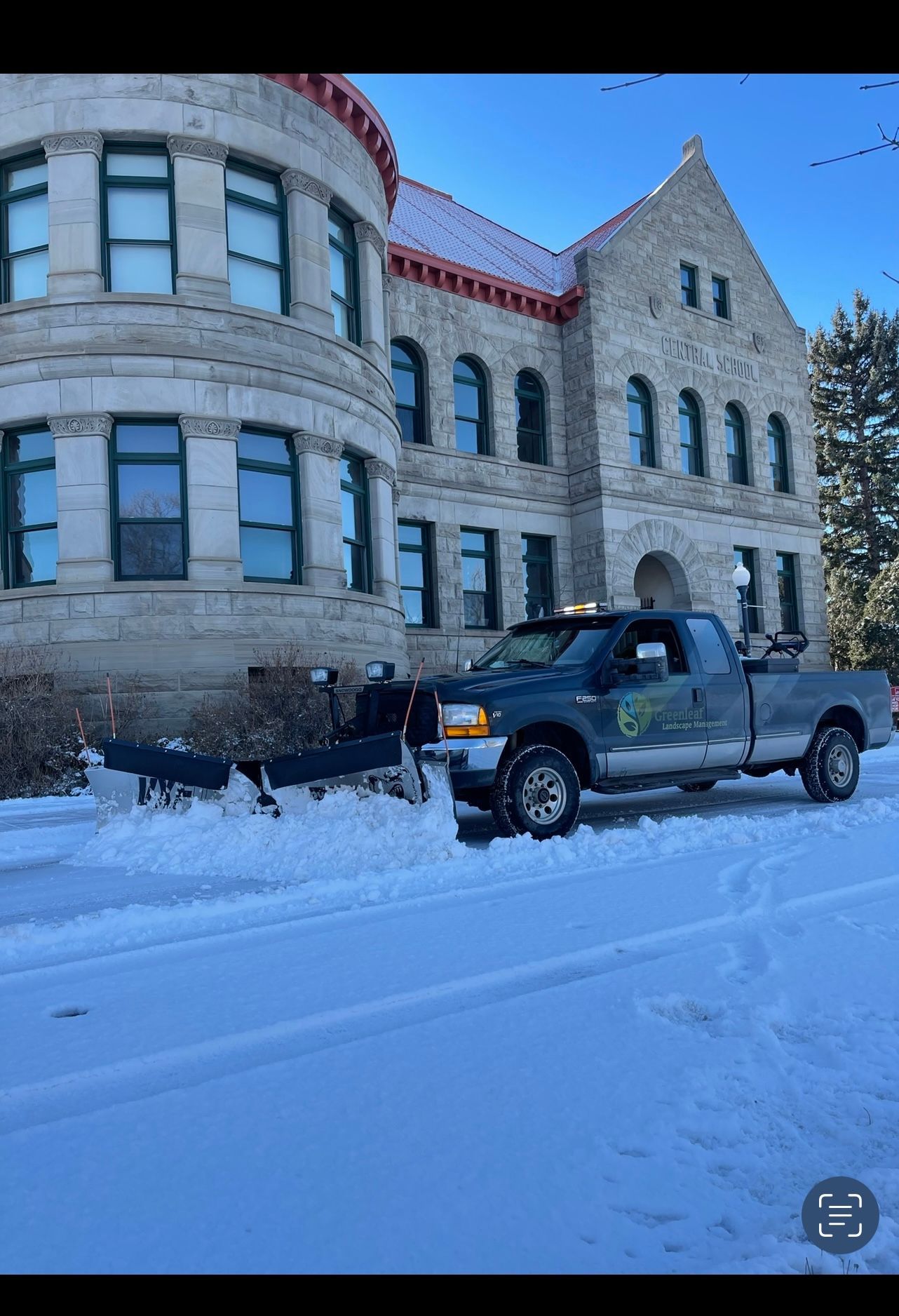 Snow plow truck clearing a snow-covered road in front of a stone building on a sunny day.