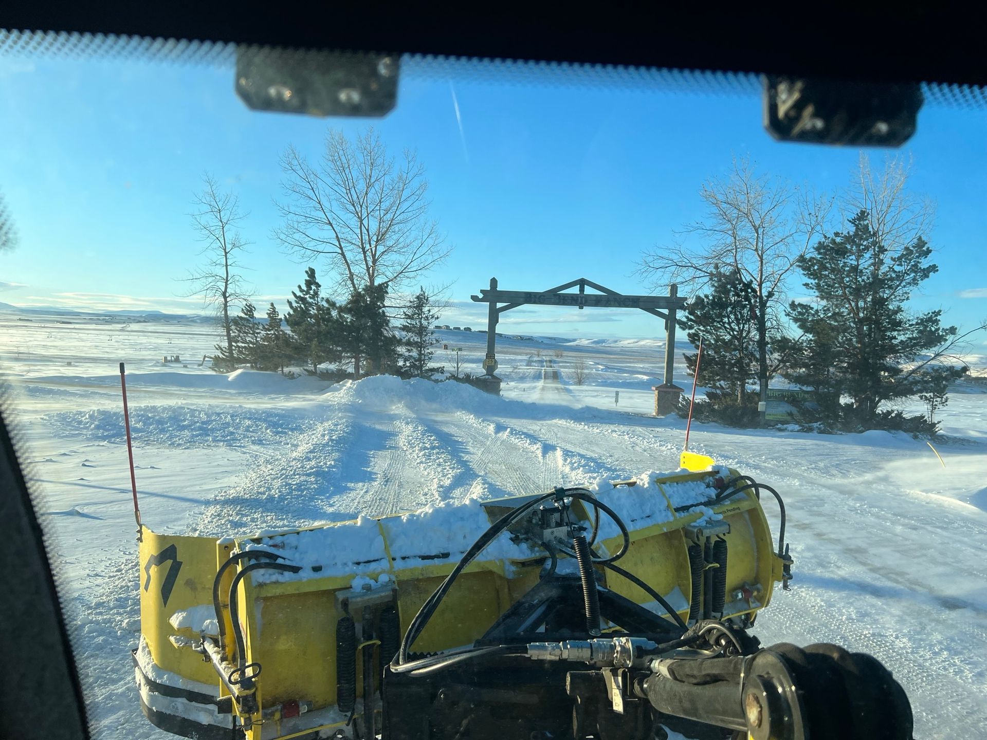 A snowplow clearing a snowy driveway towards a wooden gate under a blue sky.