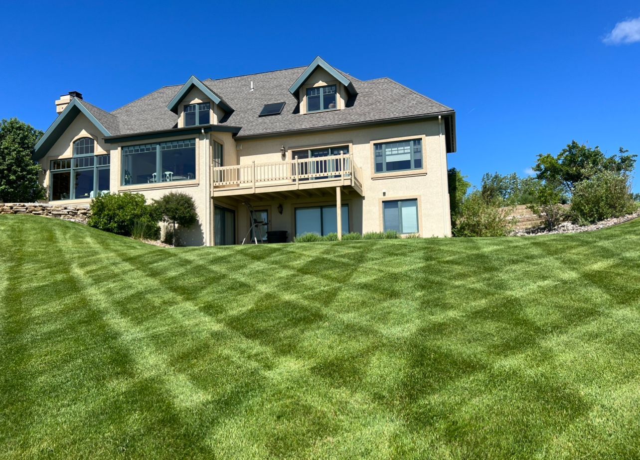 House on a hill with a green lawn mowed in a checkerboard pattern, against a bright blue sky.