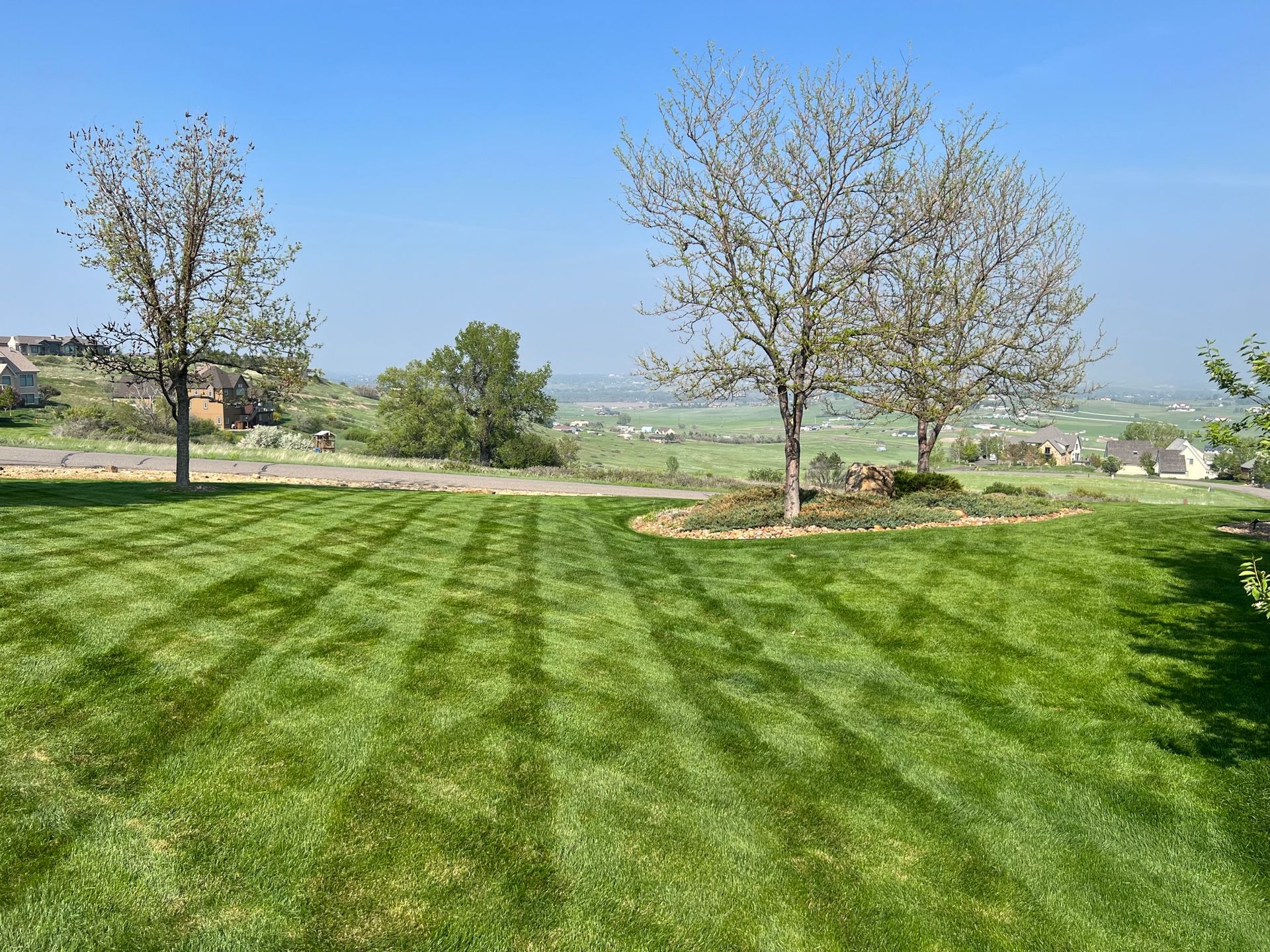 Lawn with freshly cut stripes, trees, and a distant view of a valley under a clear blue sky.
