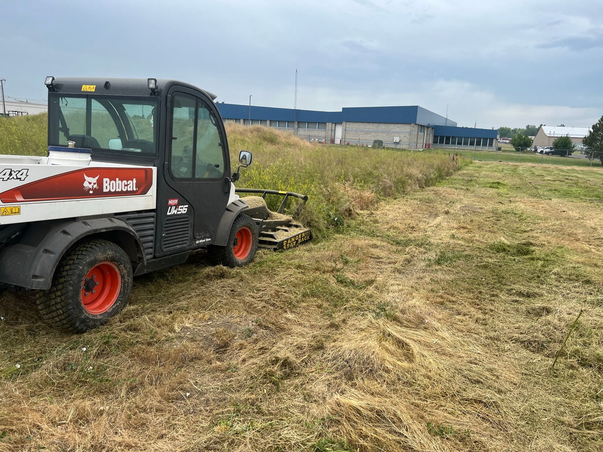 A Bobcat mower cutting tall grass in a field, with a blue building in the background.