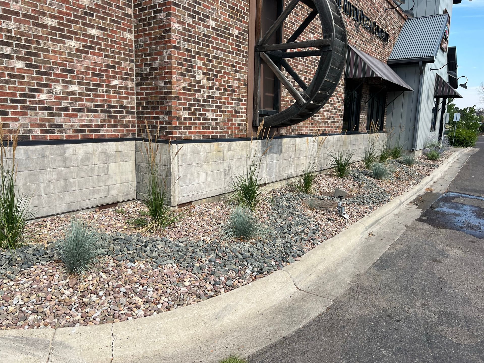 Brick building with water wheel, planting bed with gray gravel, plants, and concrete curb.