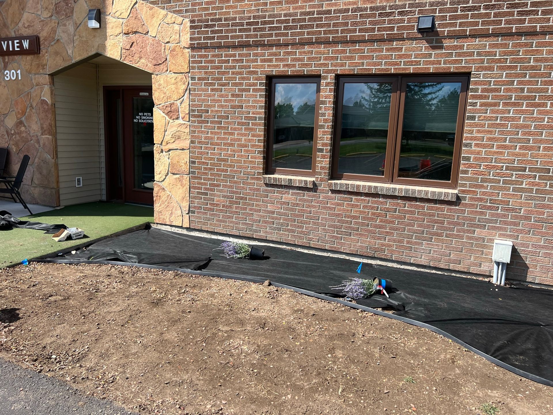 Building exterior with new landscaping. Brick wall, windows, and a gravel bed with weed barrier are visible.