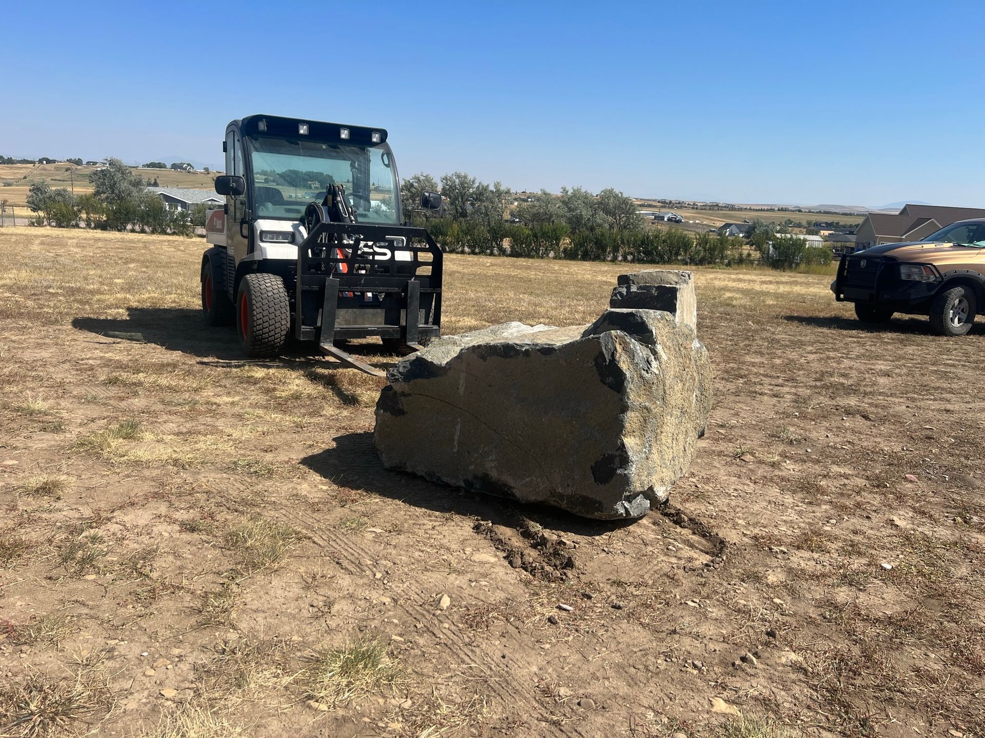 Skid steer moving a large rock across a dry field on a sunny day.