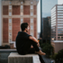 Photo of a man wearing black with glasses sitting on a rooftop looking out to a urban landscape.